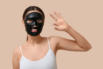Young woman with activated charcoal mask on her face showing OK against beige background