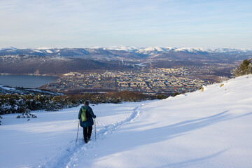 A woman with trekking poles and a backpack walks along a snowy mountain slope. In the distance is a large northern city. Path in the snow. Magadan, Magadan region, Russia. Travel to the Russia.