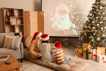 Happy family in Santa hats with popcorn watching Christmas movie on projector screen at home, back...