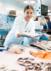 Responsive shop assistant keeping in hands fresh seabass in fish store with big assortment