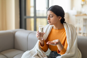 Concerned indian woman wrapped in blanket holding glass of water and medicine