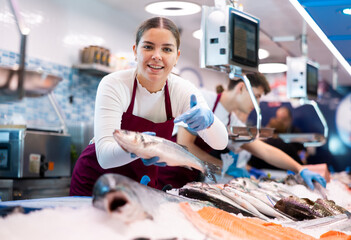 Positive young saleswoman demonstrating seabass to client in fish store with large stock - obrazy, fototapety, plakaty