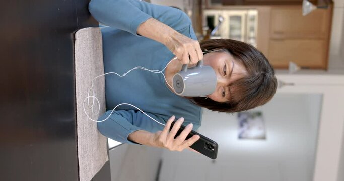 Beautiful woman relaxing drinking coffee wearing ear buds enjoying listening to music on mobile cell phone sitting at home vertical. 