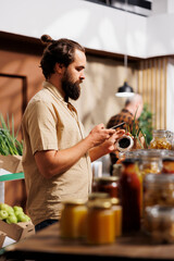 Man in zero waste store using mobile phone camera to analyze products. Vegan customer thoroughly checking local supermarket food items are suitable for his diet, using internet