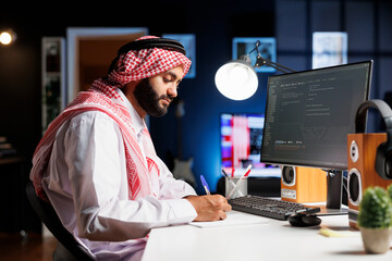 Muslim man sits at a computer and programs, exhibiting the app development process. Side-view image...