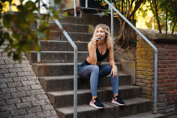Smilling fitness woman in sportswear eating fitness bar while sitting on stairs outdoors. Blonde...