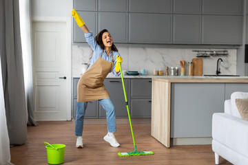 Joyful woman singing with mop like microphone in kitchen