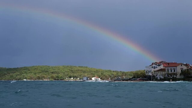 Fast motion colourful rainbow above the disquiet sea surface. Continuously changing weather conditions, light and shadow. Powerful waves crashing the coast after Storm Ciar&aacute;n smashed into Krk Island.