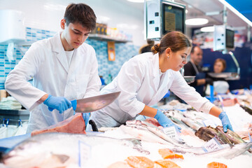 Proficient sellers in white work clothes processing tuna on table board in fish shop - obrazy, fototapety, plakaty