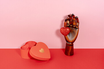  St Valentine's still life with half open red with golden detail heart shaped box with petals inside and brown wooden mannequin hand holding sparkling heart on pink and red background