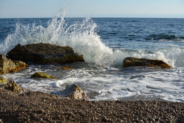 The wave breaks beautifully on the rocks on the shore, seascape, sunny day, rocks, blue sea, sea foam, vacation in the south, horizontal photo