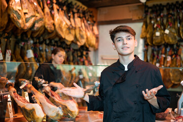 Positive young guy seller in black jacket standing near counter in butcher shop, selling dry-cured spanish jamon