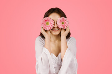 Cheerful young woman with a playful expression covering her eyes with two pink gerbera flowers