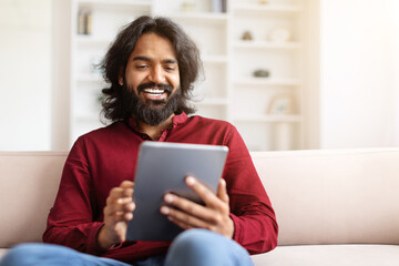 Relaxed eastern guy scrolling, using digital tablet at home