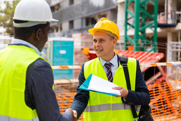 Two successful civil engineers at a construction site exchange a friendly handshake, holding...