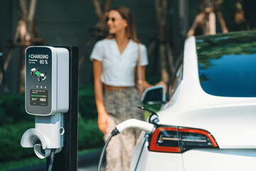 Young woman travel with EV electric car charging in green sustainable city outdoor garden in...