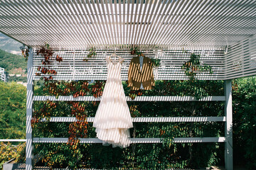 Bride dress and groom suit hang on hangers under a slatted canopy in the garden