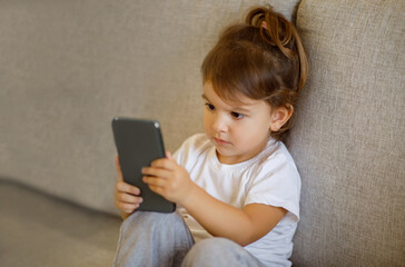 Curious toddler girl using smartphone, sitting on couch in living room