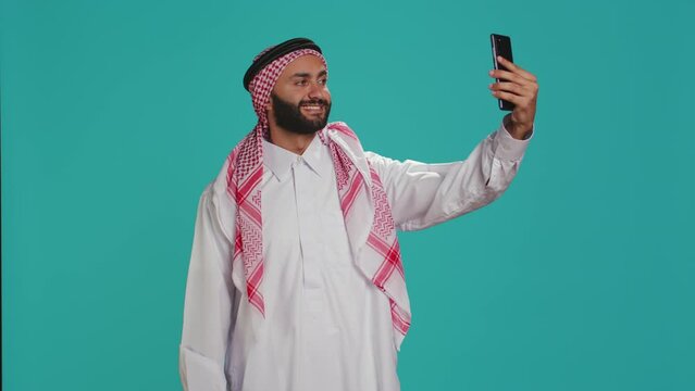 Young adult taking photos with phone, capturing memories and funny faces on smartphone camera over blue background. Middle eastern guy with checkered headscarf takes pictures in traditional attire.