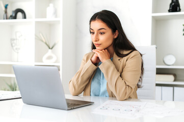 Serious concentrated young adult indian or arabian business lady, executive, company employee, lawyer, in formal wear, sitting in modern office, focused looks at laptop screen, studying information