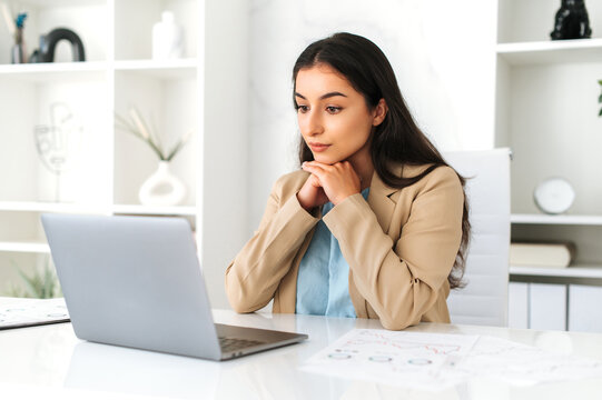 Serious concentrated young adult indian or arabian business lady, executive, company employee, lawyer, in formal wear, sitting in modern office, focused looks at laptop screen, studying information