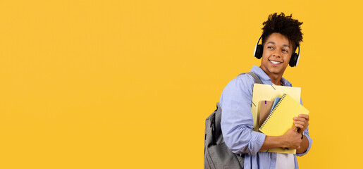 Smiling black male student with headphones and notebooks on yellow - obrazy, fototapety, plakaty
