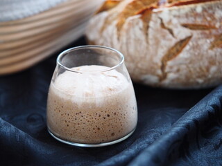 Closeup on fresh sourdough starter in a glass jar, loaf and banneton behind, black tablecloth