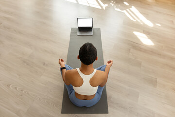 A woman in a meditative yoga pose sits in front of a laptop on a mat