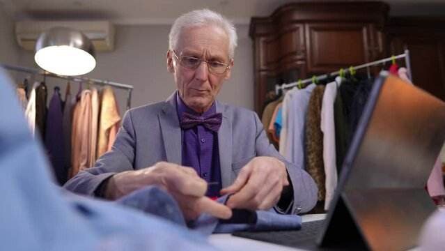 An elderly fashion designer wearing glasses is sitting and working at a table in the workshop room of his house. The man takes a pin and fixed the gathered fabric on the blue shirt.