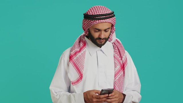 Muslim guy holding smartphone and feeling pleased after hearing good news, typing online messages on social media network. Middle eastern person scrolling internet pages, traditional clothes.