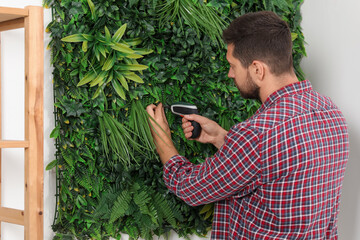 Man with screwdriver installing green artificial plant panel on white wall in room