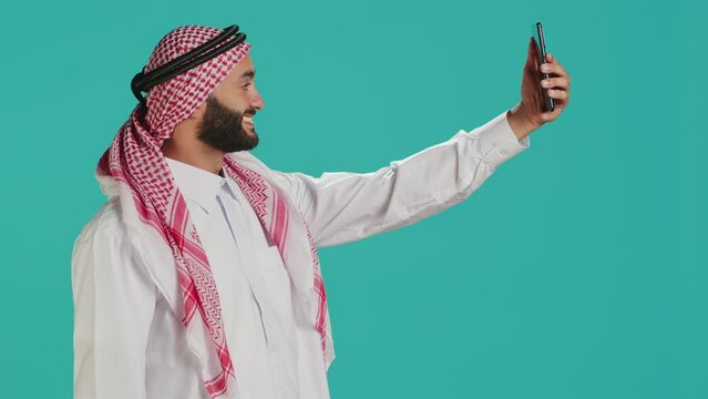 Young arab person takes photos with phone, capturing funny moments on smartphone camera in studio. Middle eastern guy with national headscarf taking pictures in traditional attire.