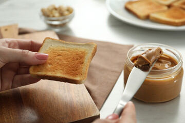 Woman spreading tasty nut butter onto toast at white table, closeup