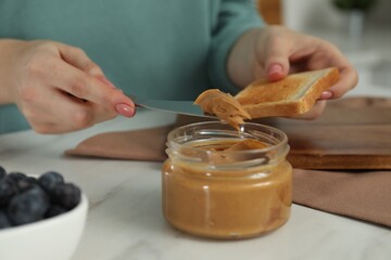 Woman spreading tasty nut butter onto toast at white table, closeup