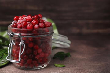 Frozen red cranberries in glass jar and green leaves on brown textured table, closeup. Space for text