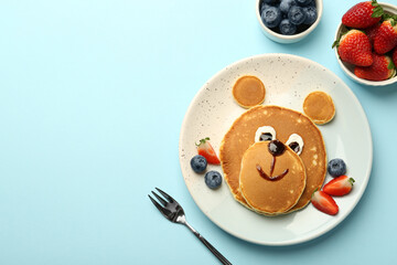Creative serving for kids. Plate with cute bear made of pancakes and berries on light blue table,...