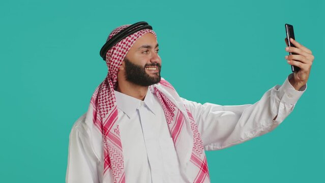 Young muslim guy takes pictures with phone, capturing important things on smartphone camera in studio. Middle eastern person with headscarf taking selfies wearing traditional arab attire.