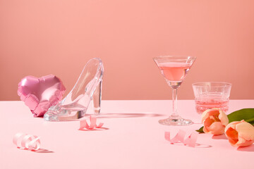 A heart-shaped balloon, glass high heel, tulip flowers and wine glasses arranged on pink surface....