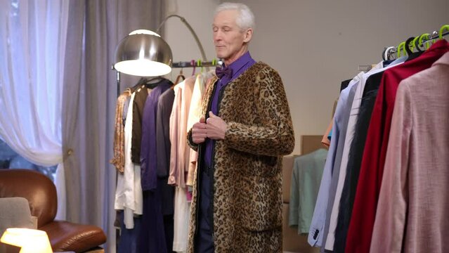 An extravagant elderly male fashion designer tries on a stylish leopard print coat, standing in the dressing room of a modern house