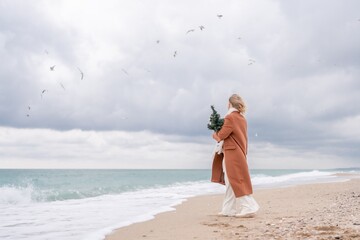 Fototapeta na wymiar Blond woman Christmas tree sea. Christmas portrait of a happy woman walking along the beach and holding a Christmas tree on her shoulder. She is wearing a brown coat and a white suit.