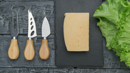A large piece of cheese, lettuce leaves and cheese knives on a wooden table.