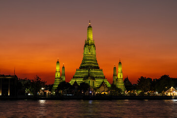 Fototapeta premium Wat Arun (Temple of the dawn) in Bangkok at dusk. Temple and prangs bathed in yellow light. Deepening orange sky in background. Chao Phraya river in foreground. 
