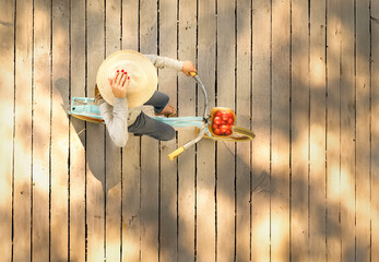 Above shot of a woman riding a bicycle carrying tomatoes on a wood pier wearing a straw hat. There...