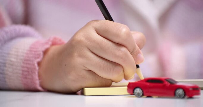 Close-up of a hand holding a pencil and writing something on a yellow paper, behind a red toy car that signifies car loan financing.