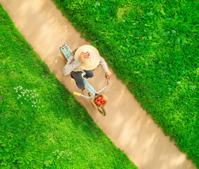 Above and diagonal shot of a woman riding a bicycle on a dirt road carying tomatoes and wearing a straw hat . There is green grass on either side of the dirt pathway.