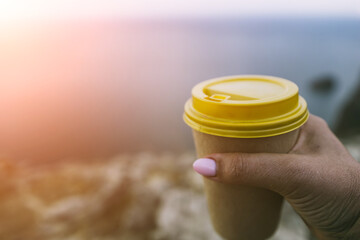 Hand holding Yellow cup with lid, coffee against a backdrop of a blue sky and sea. Illustrating cup...