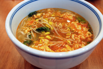 Close up of a porcelain bowl of egg tomato noodle soup on a table. Ingredients also include green vegetables and bean sprouts. A popular traditional noodle dish from China
