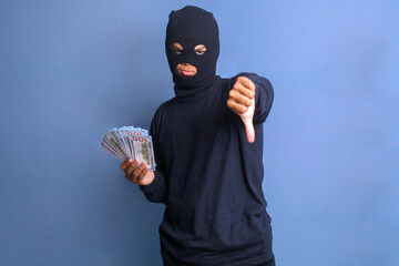 Bank robber wearing black balaclava for disguise, holding money while giving thumb down showing not satisfied expression