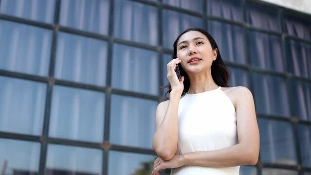 A confident Asian businesswoman stands talking on the phone with her business partner in the office outside outdoors using an application on a mobile phone fast connection online communication concept