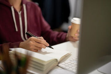 Cropped image of a man sitting in front of a computer, taking notes in a book, working in a room.